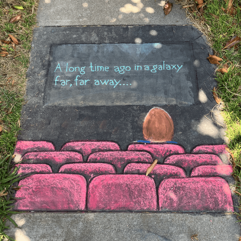 A chalk drawing on pavement featuring a darkened movie theatre with red seats; one audience member is seated, with red hair; on the screen, "A long time ago in a galaxy far, far away" is written in blue lettering.