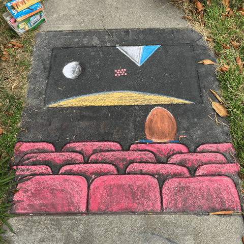 A chalk drawing on pavement featuring a darkened movie theatre with red seats; one audience member is seated, with red hair; on the screen, the opening scene from the first Star Wars movie, with a planet, a moon, and two space ships