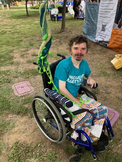 A young man with light skin and dark hair and goatee, wearing a blue t-shirt and sitting in a wheelchair outdoors. A crocheted beanstalk is attached to the back of his chair.