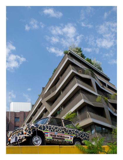 Low shot taken looking upwards toward (in the background) a high-rise block of flats seen diagonally and (in the foreground) an old "Trabi" car in leopard skin colours mounted on a pedestal above the viewer.