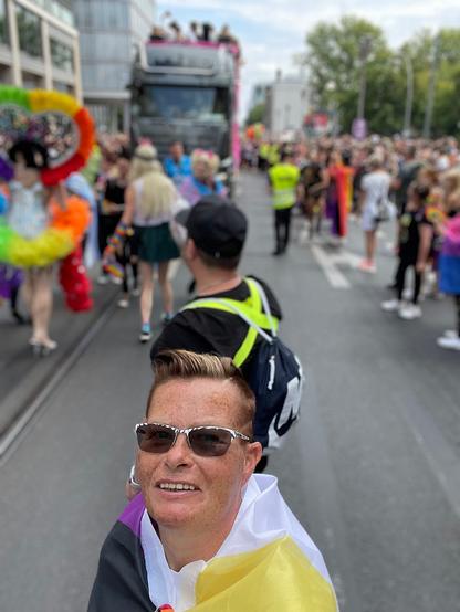A person with short reddish hair and sunglasses, wrapped in a nonbinary pride flag, smiles at the camera during CSD Berlin 2022. Behind them, a colorful and lively crowd marches down a city street, with floats, rainbow outfits, and celebration all around.