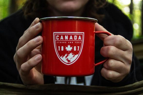 A close-up of two hands cradling a red tin mug that says, “Canada — Strong and Free — 1867” and has a stylized picture of mountains and a moose.