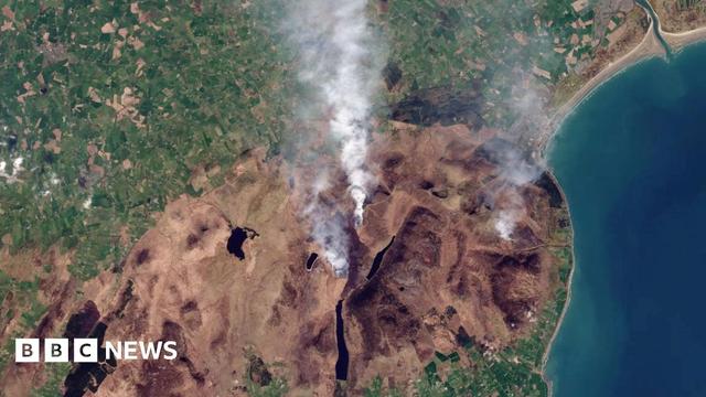 A satellite image shows a large area of brown vegetation at its centre, with some smoke visible. Green, unburned agricultural fields are visible to the north. The coastline and sea can be seen to the east.