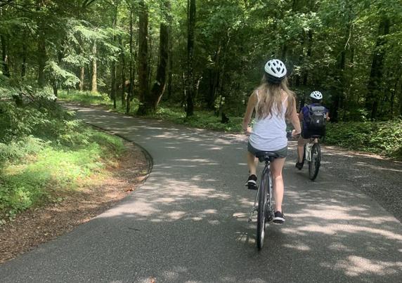 Two bikers with helmets biking on a one lane road through the forest. One biker has a backpack.