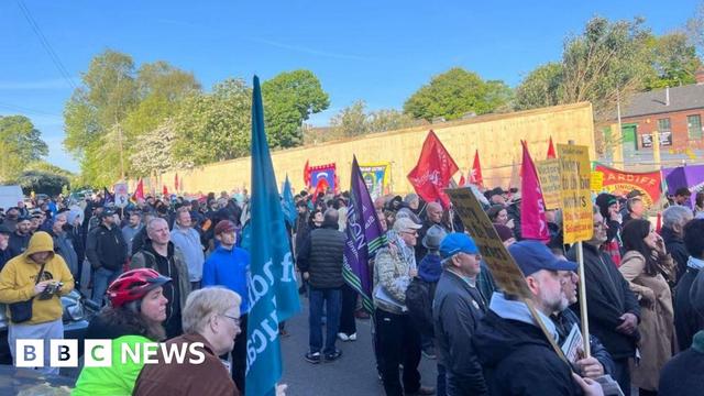 A crowded street, filled with people protesting against the Birmingham bin strikes. They are holding flags and placards. The sky is bright blue in the background.