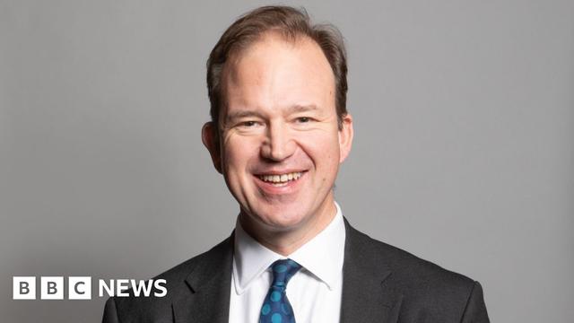 A portrait photograph of MP Jesse Norman. He is wearing a grey suit jacket and white shirt with a blue spotty tie. He is stood against a grey backdrop.