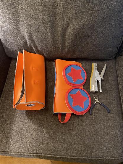 Orange, blue, and red vinyl bag pieces being assembled into a star tube duffel shaped purses. Plier stapler and blue handles pliers.