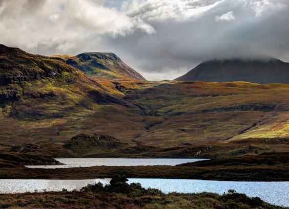 A striking landscape image of the essence of remote highland Scotland, featuring wild mountains and loch water in the foreground. The location is famous and loved for its ever changing at speed, play of shafts of brilliant sunlight through cloud, suddenly illuminating like a spotlight on features of the raw land, a vision which has inspired so many writers and artists.