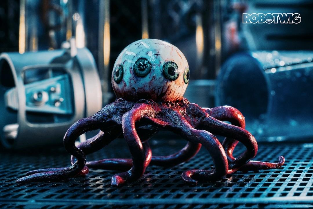 The Eye escapes from its container (tipped over in the background) and walks along a work surface. It has four pupils on show and many tentacles at its base. The Eye escapes from its container (tipped over in the background) and walks along a work surface. It has four pupils on show and many tentacles at its base.