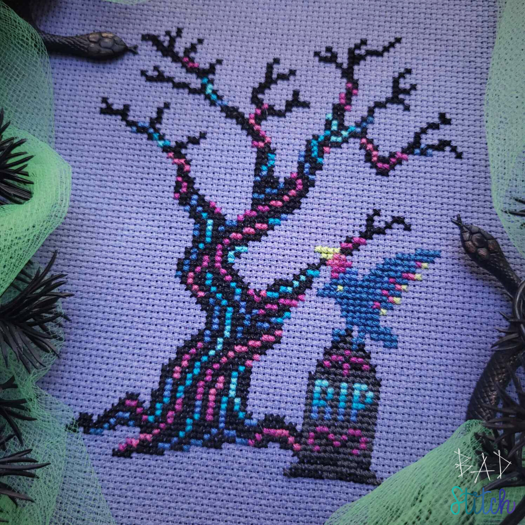 A cross stitch on light purple fabric, staged with neon green tulle, fake foliage, and little snake figurings.  The cross stitch shows a black, gnarly tree with vaporwave lines running through the trunk and branches. A headstone sits next to the tree with RIP engraved upon the stone and a raven sits perched with wings extended.  BAD Stitch.