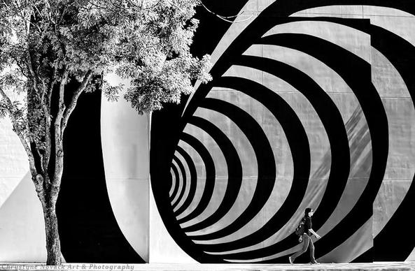 A black and white photograph capturing a moment in time. Strolling by a large op-art wall mural on the side of a building a young adult appears to be warping into another dimension.