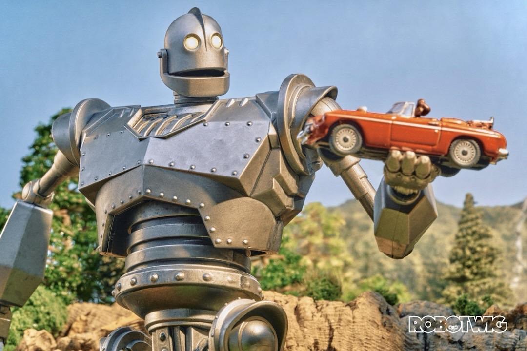 The Iron Giant holds an old red sports car with Hogarth in the drivers side ready for the Giant to spin him around in it like a theme park ride