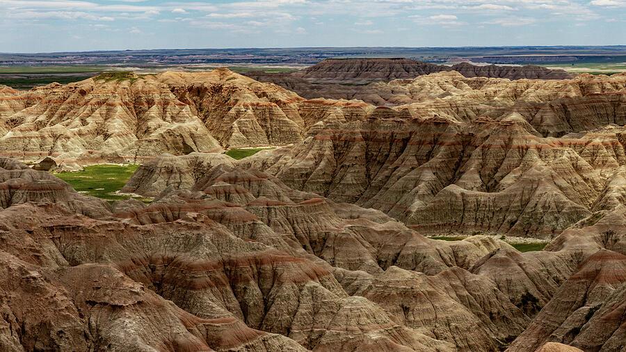 Badlands Panoramic

The landscape stretches out like a vast, ancient cathedral of stone, its sharply ridged peaks and layered sedimentary bands rising in jagged rhythms across the horizon, each ridge catching light differently as if the earth itself were breathing. 
. Under the bright blue summer sky, the formations glow with shifting tones of tan, red, and gray, textures so intricate you can almost feel the grit of wind‑carved rock beneath your fingertips. The air seems dry and sun‑warmed, carrying the quiet weight of a place shaped by centuries of erosion—wild, untamed, and humming with the timeless energy of the American West.