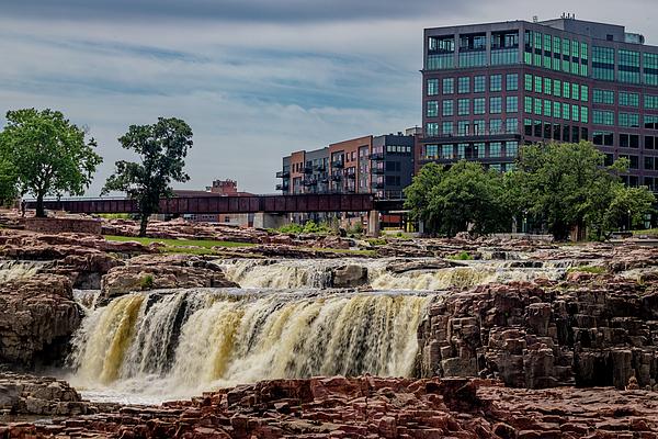 Sioux Falls South Dakota

The scene unfolds with the steady roar of Sioux Falls pouring over rugged, time‑worn rock—water churning white as it crashes into the pools below, sending a cool mist that you can almost feel on your skin. Sunlight warms the stone, revealing earthy reds and browns that contrast beautifully with the crisp, flowing ribbons of water. Behind the falls, the modern downtown skyline rises in clean lines and muted tones, adding a subtle hum of urban energy to the wild, tactile textures of the park. The whole image carries a sense of movement, freshness, and grounded Midwestern strength, where nature’s raw force meets the calm geometry of the city.