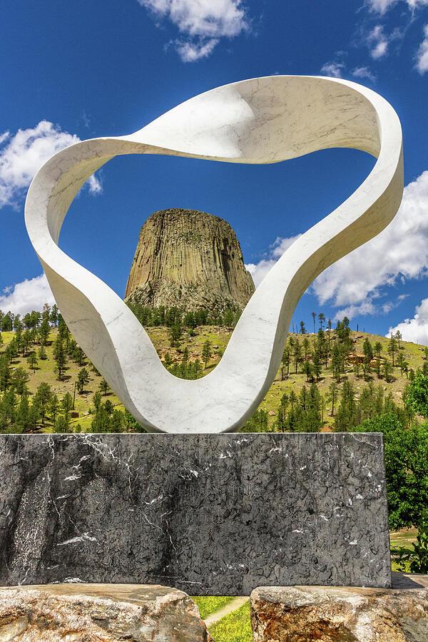 Devils Tower and the Sacred Circle of Smoke

The scene opens with a sense of stillness and ceremony. In the foreground, the Sacred Circle of Smoke sculpture rises in a smooth, looping arc—its polished surface catching the Wyoming light in a soft, silvery glow. The curve feels almost alive, like a ribbon of smoke paused mid‑ascent, and its shape naturally draws your eye through the opening toward Devils Tower, standing rugged and immense in the distance. 
The air in the image feels crisp and open, carried by a clear blue sky streaked with scattered clouds that drift like slow‑moving brushstrokes across the horizon.  The contrast between the sculpture’s smooth, contemporary form and the Tower’s weathered volcanic columns creates a tactile tension—you can almost feel the cool metal beneath your fingertips and the rough stone rising beyond it.
There’s a quiet spiritual energy woven into the composition. The sculpture’s symbolic gesture—honoring the rising smoke of prayer and ceremony—adds a sense of reverence, as though the landscape itself is participating in something sacred and ongoing. The framing of Devils Tower within the circular opening feels intentional and ceremonial, linking modern artistry with Indigenous tradition in a way that deepens the emotional weight of the scene.