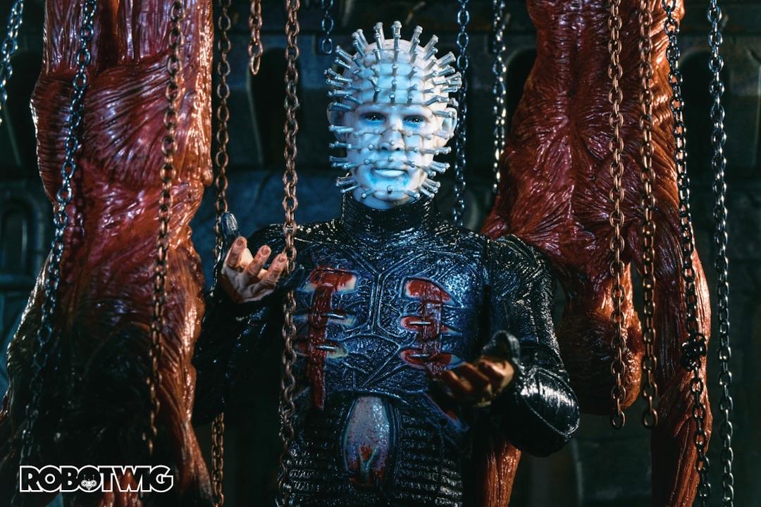 Pinhead stands amongst hanging corpses and chains inviting you to join him