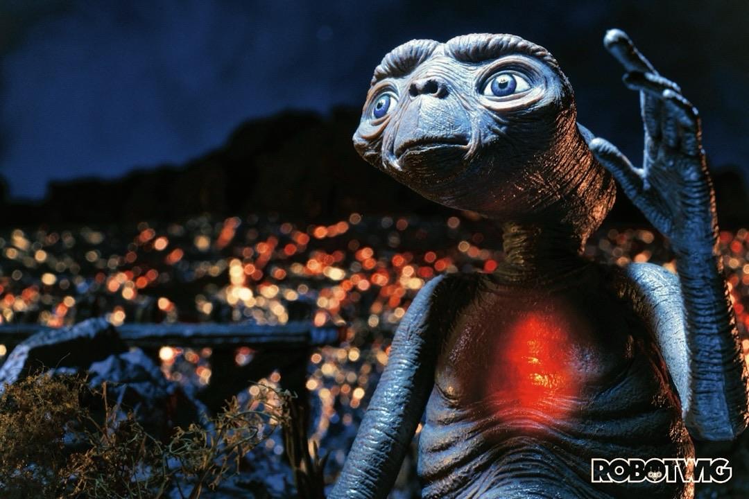 E.T. Looks up at his ship leaving as he waves his arm to get their attention. His chest glows brightly. City lights can be seen behind him