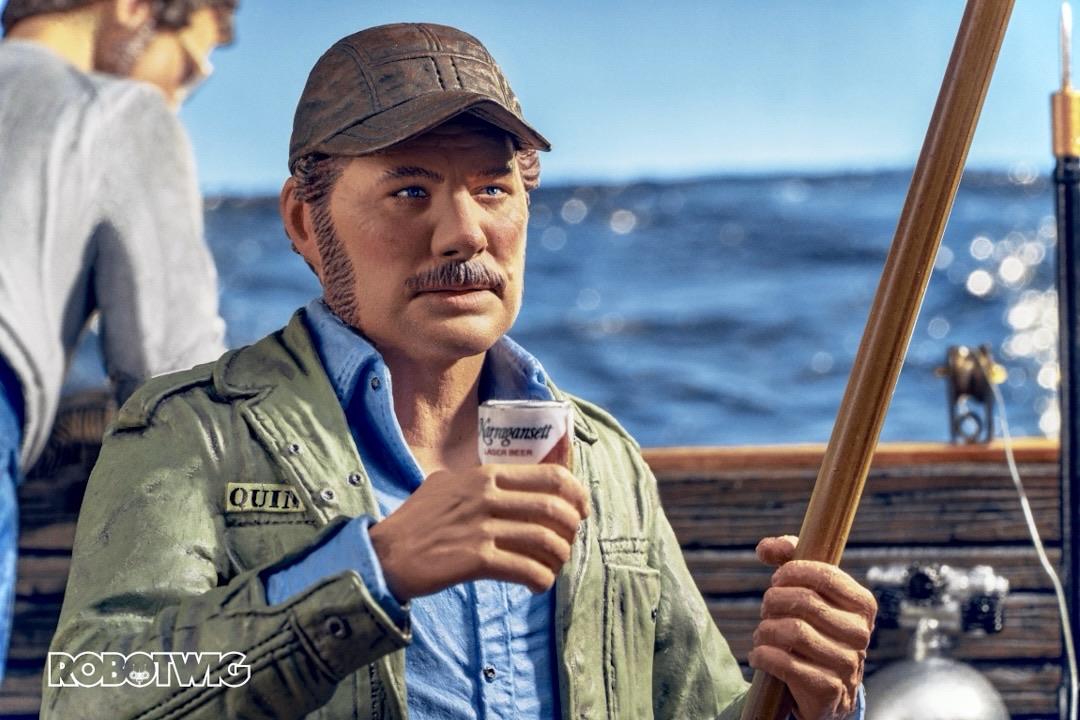 Quint holds a beer in one hand and a fishing rod in the other whilst sat down on the orca telling one of his stories