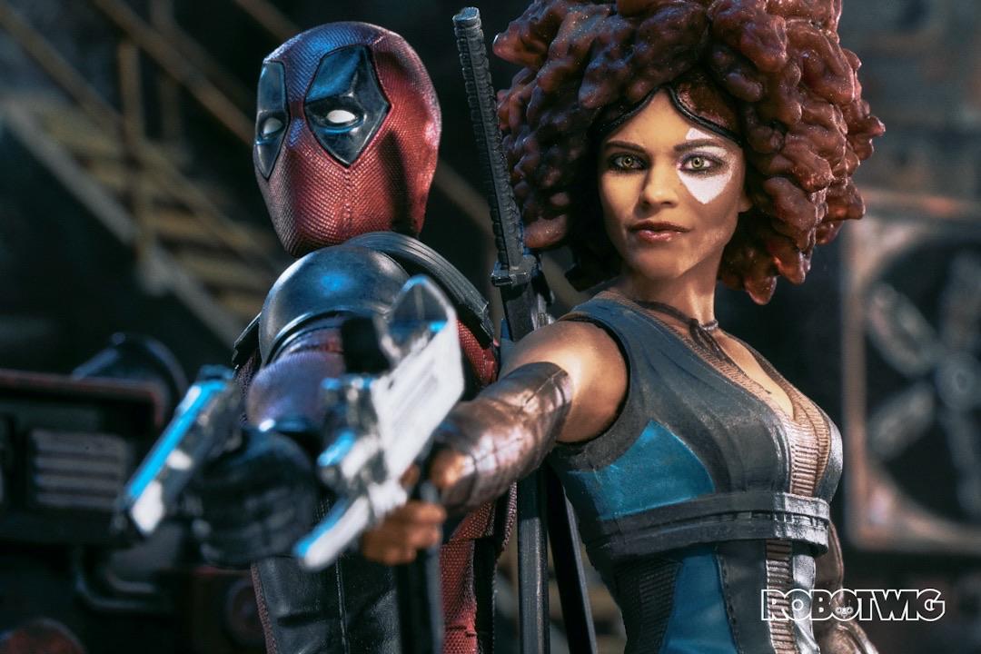Deadpool and Domino stand in an old warehouse pointing their guns down at someone off camera
