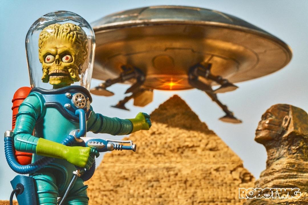 A Martian looks off camera to his other Martian friends and points over at a UFO hovering over the pyramids. A red glow comes from under the UFO ready to destroy the pyramids