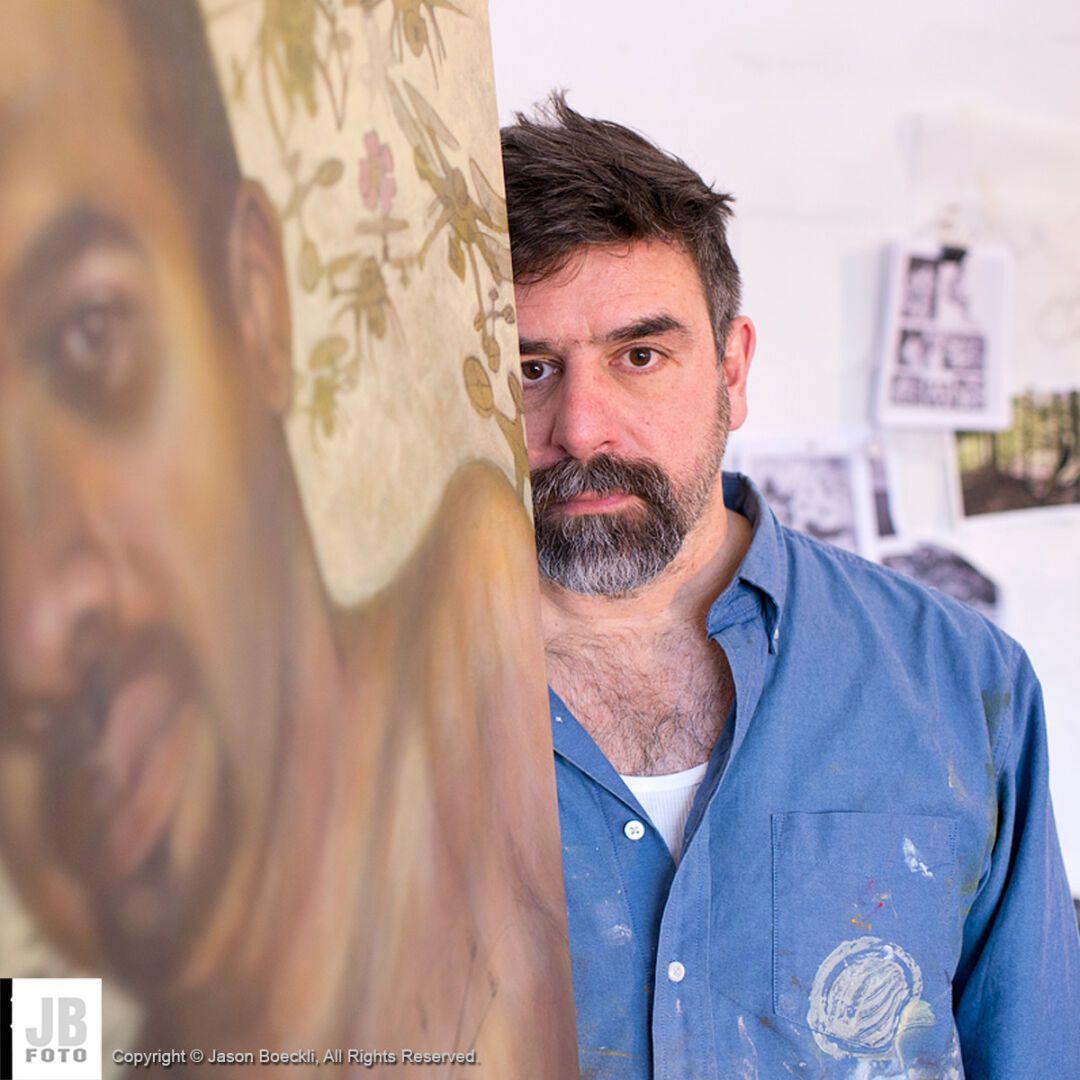 Artist Joe Radoccia standing behind a painting he was working on. He is in a blue shirt and his eyes are looking right into the viewer.