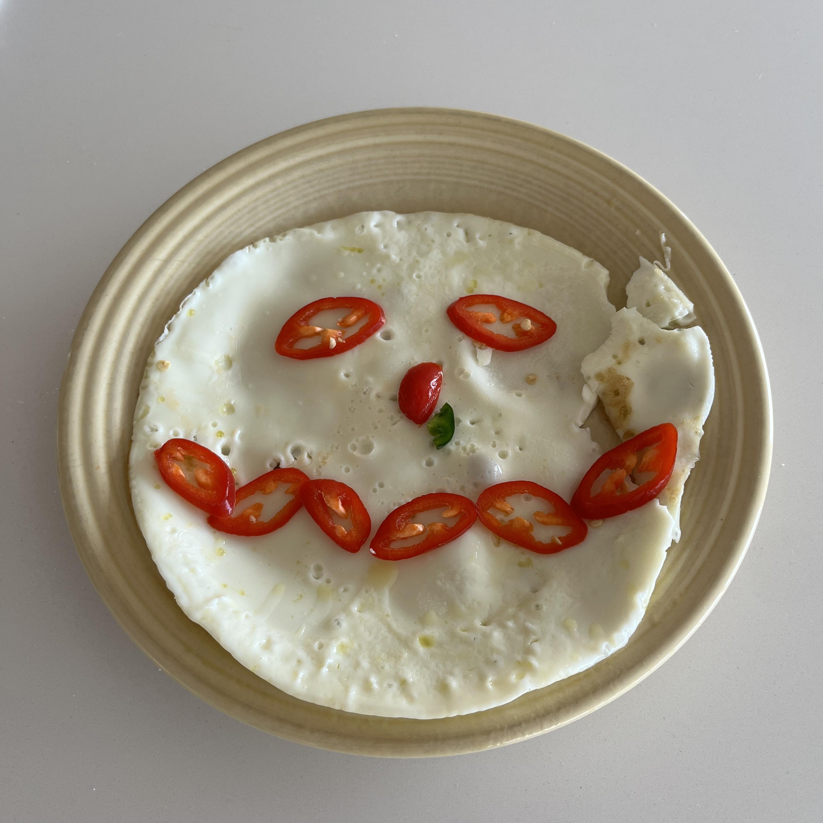 On a yellow plate, a circle of cooked egg white is garnished with red jalapeno slices in the form of a face or snowman head. The slices are all red with the exception of one small green nugget coming out of what would be the snowman's nose.