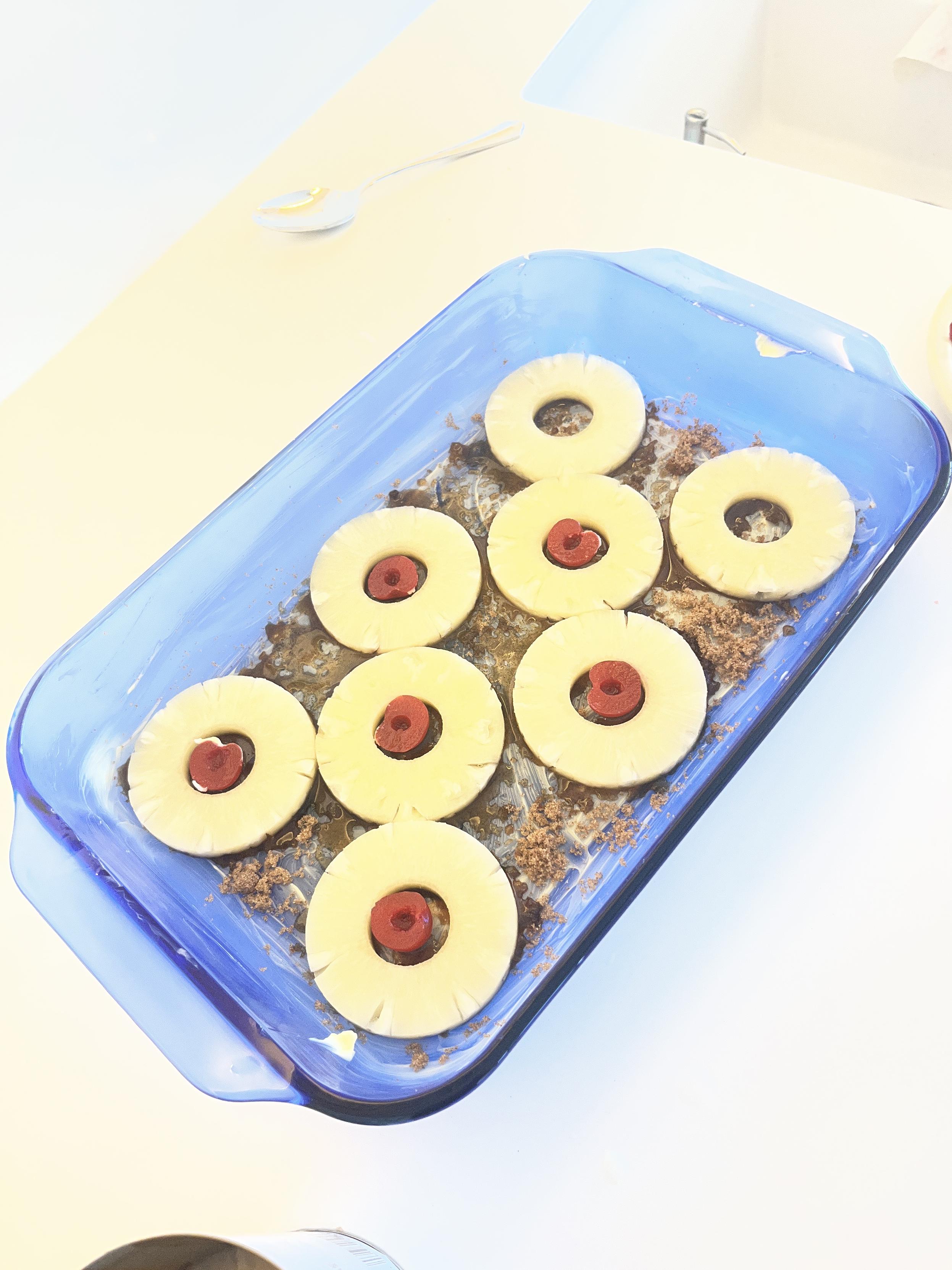 A blue glass baking pan. It is lined with butter, sprinkled with brown sugar and has an arrangement of pineapple slices and bright red maraschino cherries