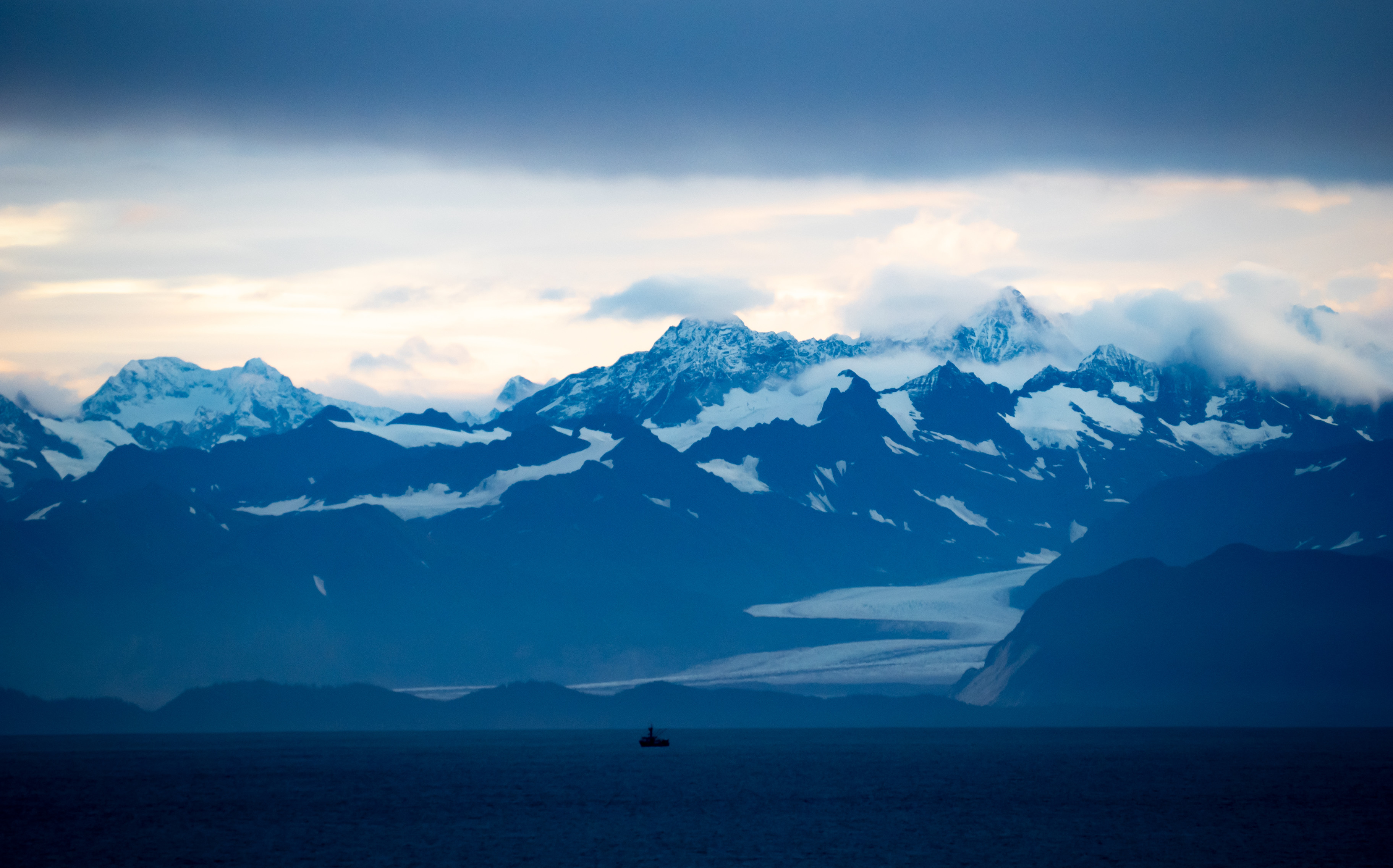 In shades of pre-dawn blue, a craggy range of mountains topped with snow and clouds, a glacier winding down to the sea at the base, where a small fishing boat is  in black silhouette