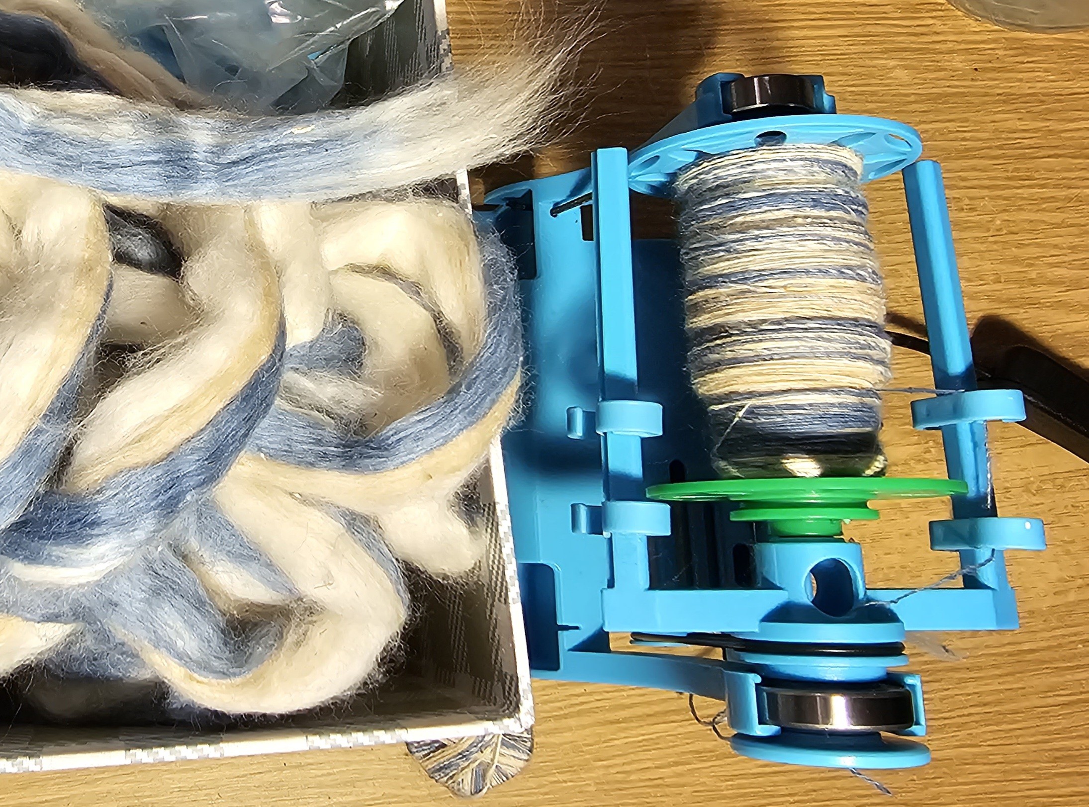 Blue and white cotton sliver in a box, next to a little turquoise plastic e-spinner, its bobbin half full