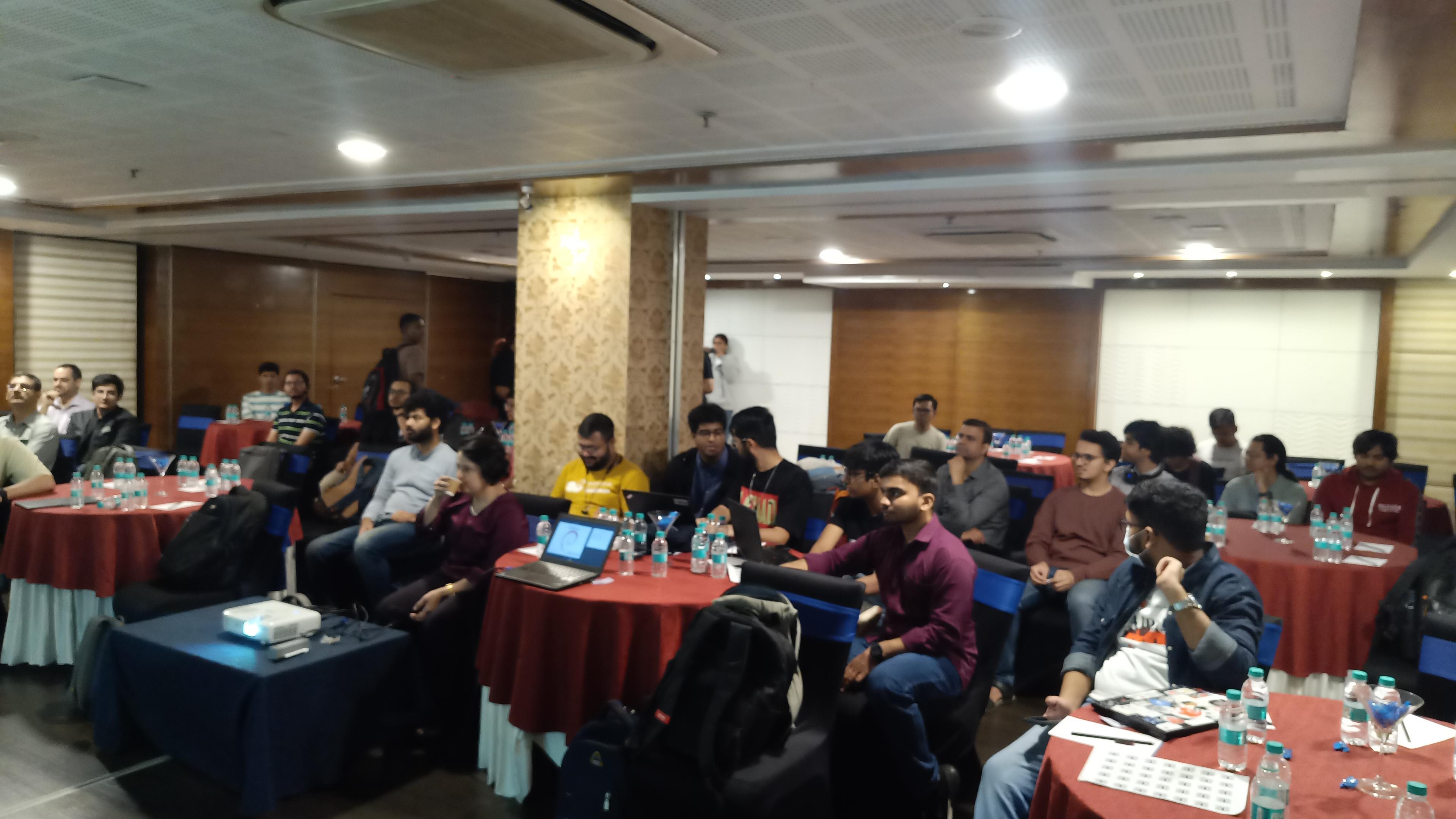 A wide shot shows a large room filled with people seated at red-clothed tables. The room appears to be a conference or meeting hall, with dark wood paneling along the walls and a large, white, blank projection screen at the front. Most of the people are facing forward, presumably toward a speaker or presentation, and many are looking at laptops or taking notes. Water bottles are visible on each table, along with some papers and other small items. The room is brightly lit by overhead fluorescent lights, and a large pillar is visible on the left side of the frame.
