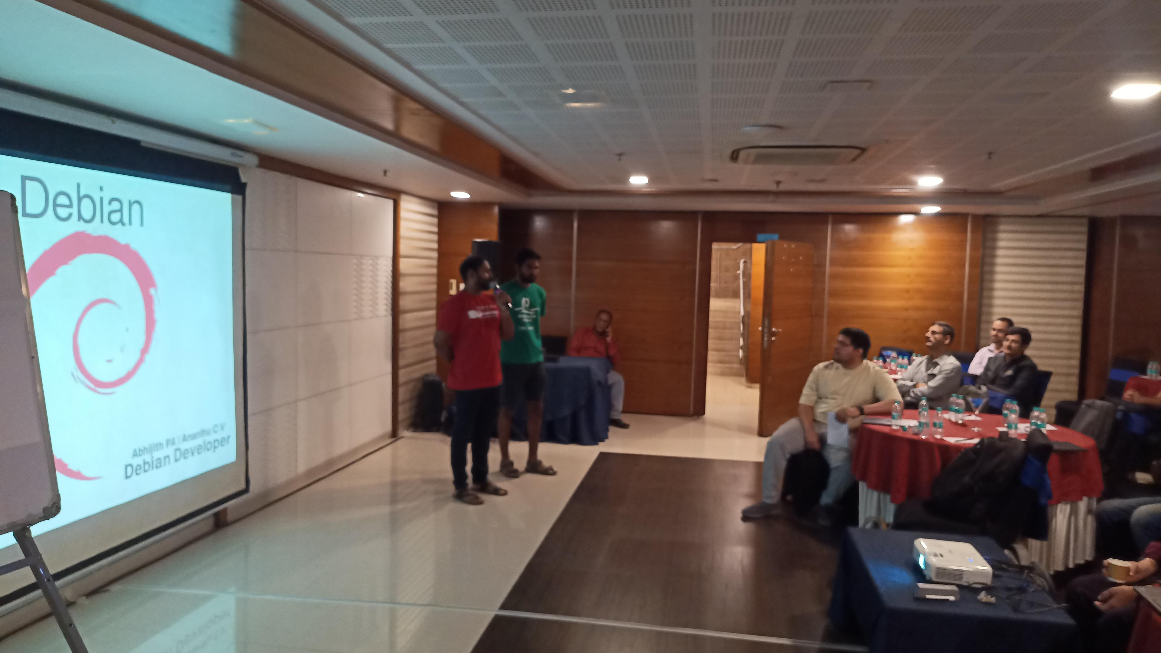 A wide-shot indoor photograph depicts a presentation or meeting in progress within a conference room. A projection screen displays the word "Debian" in large white font, with a red circular logo to the left. Two people are standing near the screen, facing a group of seated attendees. The attendees are gathered around several round tables covered with red tablecloths. The room features wood-paneled walls and a drop ceiling with recessed lighting, and a dark rug is visible on the floor. 

The text on the screen reads: "Debian". People are seated at tables with visible cups and plates. Several individuals are wearing collared shirts, and one person is wearing a bright green shirt. The room appears to be dimly lit, with the projection screen providing the primary source of illumination.