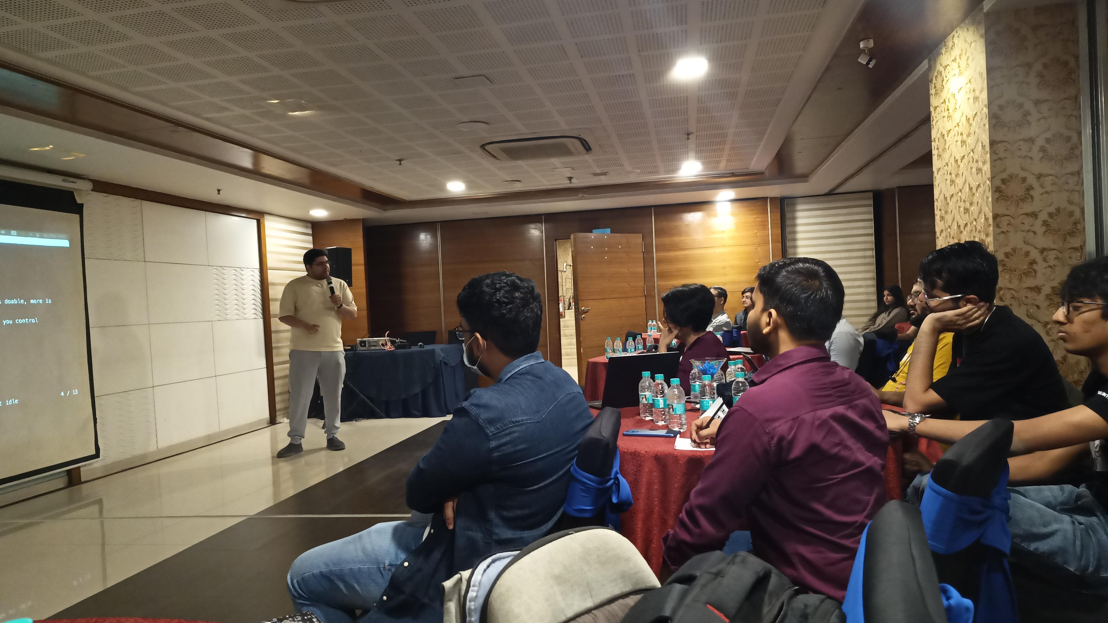  A group of people are gathered in a room for a presentation or lecture. A person stands in front of a screen, seemingly speaking to the audience. The audience members are seated in rows of chairs facing the speaker, with several tables in front of them. Most people are looking toward the speaker.  

The room has brown walls with vertical panels. There are bottles of water on the tables. A small portion of code is visible on the screen reading "text: 'This is just a test sentence'". The people in the audience are dressed casually and some have bags next to them.