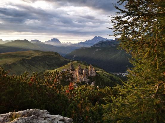 A panoramic view of a mountainous landscape featuring layers of rolling hills, rocky formations, and dense forests under a partly cloudy sky. In the foreground, green coniferous trees and a rocky outcrop are visible on the bottom left. Midground includes a hill with jagged rock pillars and scattered trees, with a small settlement in the valley below. Distant mountains rise in the background, partially obscured by clouds, under varying light suggesting late afternoon. The sky contains thick, gray clouds with patches of lighter sky, indicating overcast conditions.