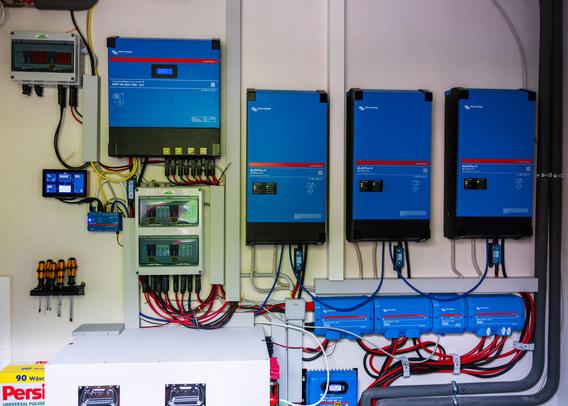 Technikwand der DIY-Photovoltaikanlage: Drei blaue Victron MultiPlus-II Wechselrichter nebeneinander, darüber ein Victron MPPT RS 450/200 Solarladeregler, Lynx Distributor, Sicherungskästen und ein kleiner Kontrollbildschirm.