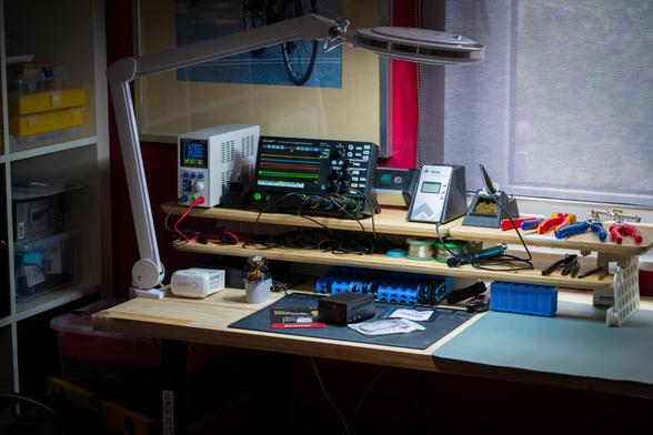 Aufgeräumter Elektronik-Arbeitsplatz mit Holzoberfläche und Lupenleuchte. Auf einer Erhöhung stehen ein Labornetzteil, ein digitales Oszilloskop mit Signalverläufen und eine Ersa-Lötstation, daneben Zangen und Werkzeug. Auf der Arbeitsmatte liegt ein kleiner Mini-PC/Raspberry Pi mit Zubehör für das nächste Bastelprojekt.