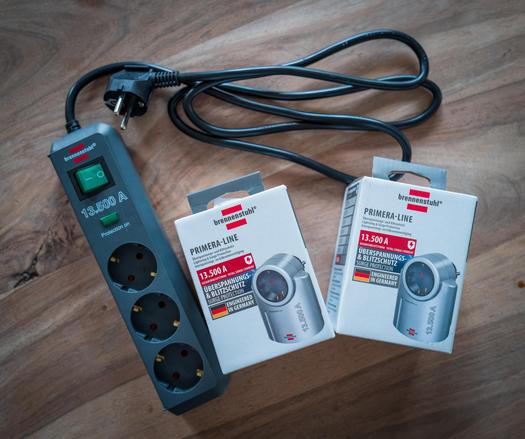 Ein kompakter, weiß-grauer Überspannungsschutz-Zwischenstecker von Brennenstuhl (Primer-Serie) liegt ausgepackt neben der Versandverpackung auf einem Holztisch.