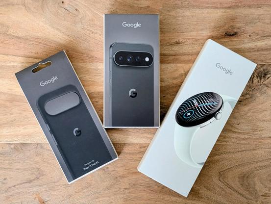 Originalverpackte Google-Produkte liegen nebeneinander auf einem hellen Holztisch.
Links eine schmale, dunkelgraue 

- Google Pixel 11 Pro XL
- Google Pixel Watch Smartwatch