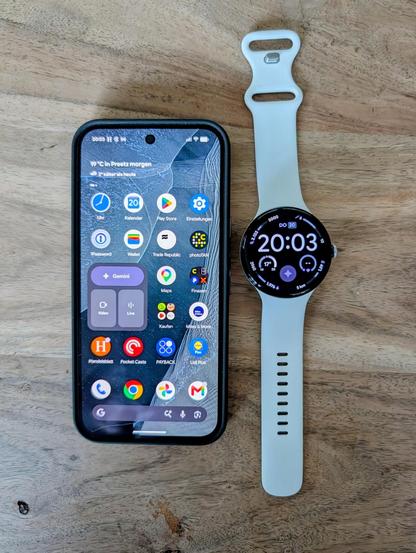 Ein schwarzes Google Pixel Smartphone in einer dunklen Schutzhülle liegt neben einer Google Pixel Watch mit weißem Silikon-Sportarmband auf einer hellen Holzoberfläche