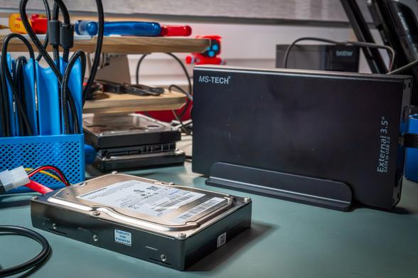 uf einer Werkbank liegt im Vordergrund eine geöffnete 3,5-Zoll-SATA-Festplatte, angeschlossen an ein Strom- und Datenkabel. Rechts daneben steht ein schwarzes, aufrecht platziertes externes Festplattengehäuse von MS-Tech („External 3.5“ SATA to USB 3.0“). Im Hintergrund stapeln sich weitere Festplatten, daneben ein blauer Werkzeughalter mit Kabeln und Schraubendrehern sowie Werkstatt- und Technikzubehör auf einem Holzregal.