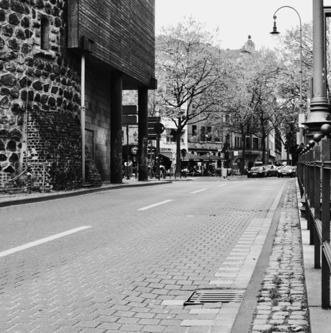 Black and white photo of a city street with cobblestone pavement, buildings, leafy trees, street lamps, parked cars, and a pedestrian walkway. There is a structure with stone wall on the left and modern architecture elements.