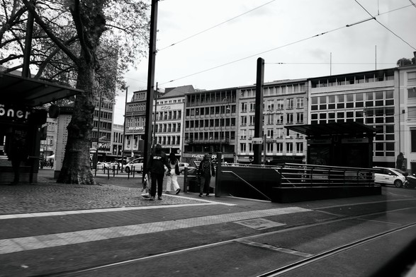 Black and white image of an urban street scene with buildings, tram lines, a pedestrian area, and people walking. There are storefronts and signs, including one for 