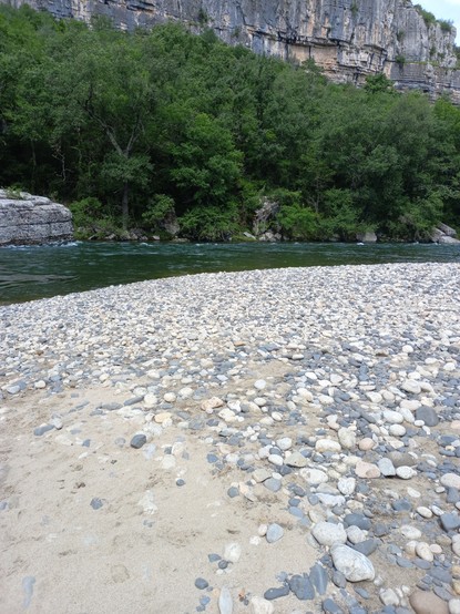 Kieselbelegter Badestrand an der Ardèche.  Gegenüber liegt ein grosser Fels im Fluss. Am gegenüberliegenden Ufer stehen Bäume und Sträucher.