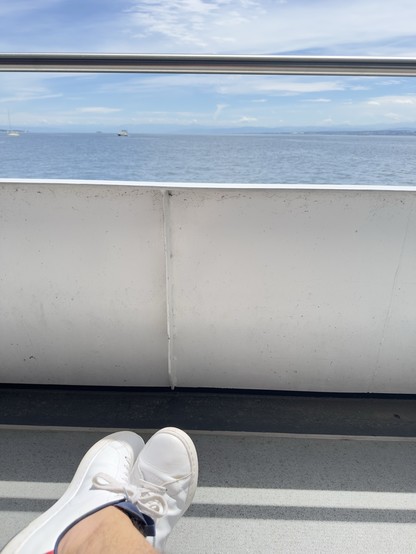 Feet in white sneakers resting on a boat with a scenic view of the ocean and distant boats under a blue sky.