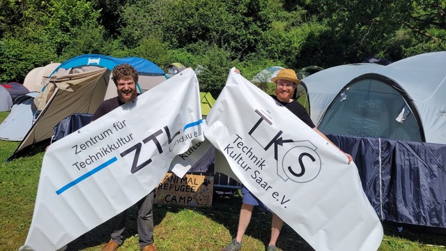 2 Personen halten ein geteiltes Banner. Eine Hälfte zeigt das ZTL Logo, die Andere das von TKS.