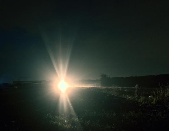 A bright light shines in a dark landscape, surrounded by a faint mist and a dimly lit field. The light appears to be coming from a vehicle or light source, illuminating the area in front of it.