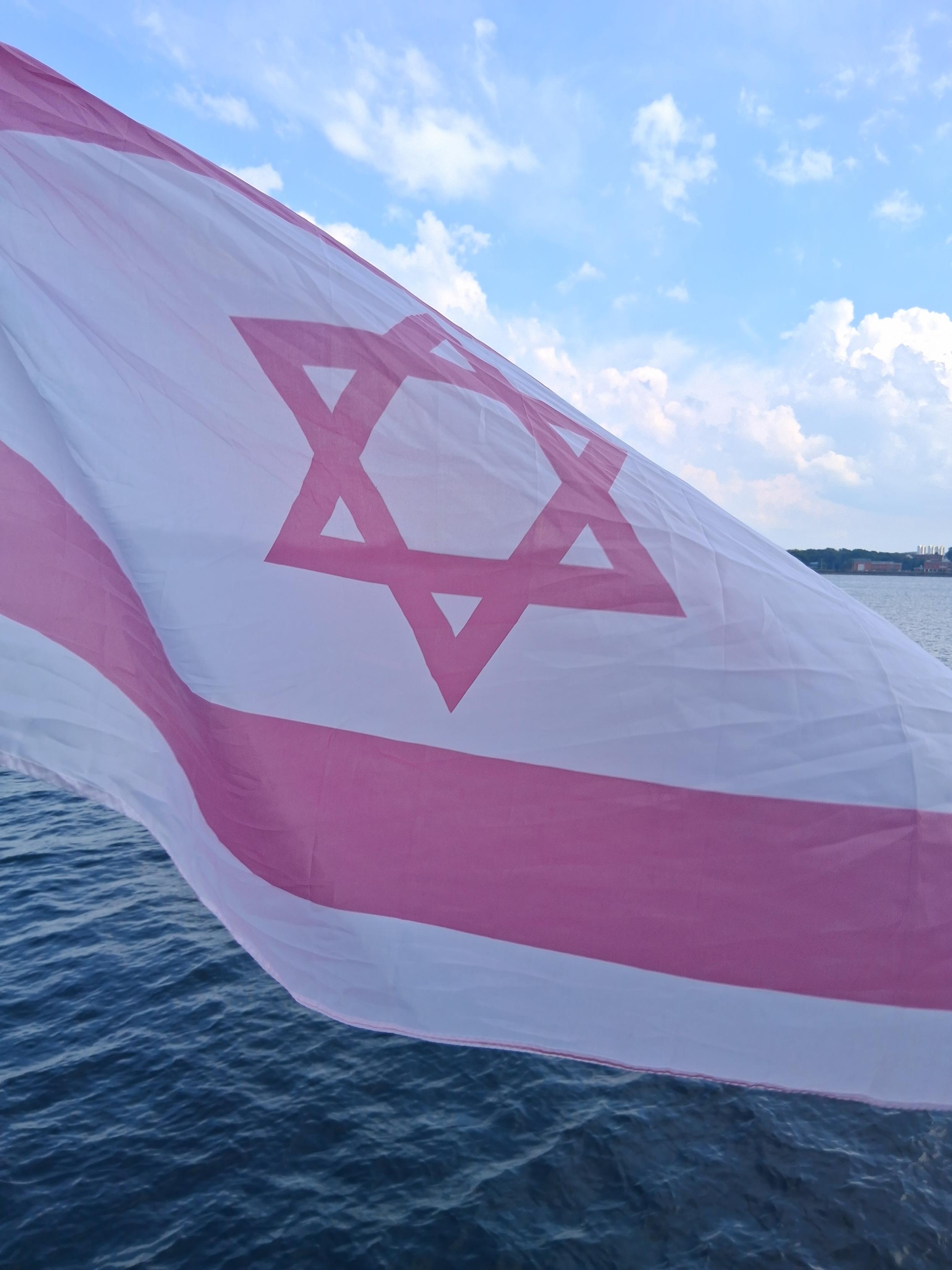 Israelische Fahne in Rosa und Weiss ( also mit rosafarbenen Davidstern zwischen zwei rosa Rechtecken). Im Hintergrund das Wasser der Kieler Förde