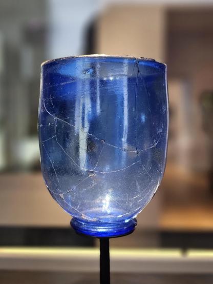 Blue translucent glass beaker, mounted on a black metal rod. The scratched and cracked surface is visible against a softly blurred indoor background.