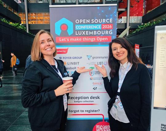 a photo showing Gina Plat (a woman with shoulder-length blond hair) and me (woman with long brown hair) smiling while standing in front of a poster reading "Open Source Conference Luxembourg 2026 - Let's Make Things Open"