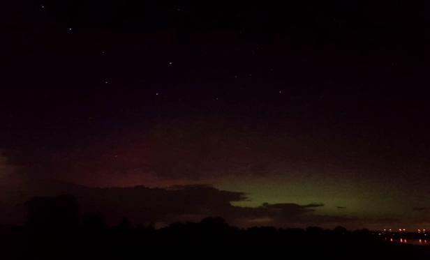 rechts die Elbe, Lichter spiegeln sich darin, der Himmel ist unten grünlich, nach oben rot von Polarlicht erleuchtet, dunkle Wolken und dunkle Baumkulisse