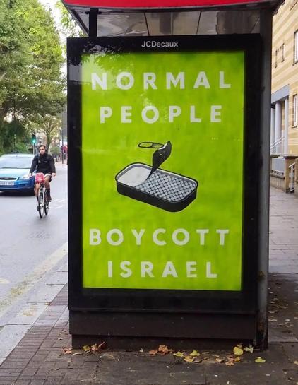 A large, bright green advertisement poster is displayed in a black JCDecaux-branded street-side frame. The poster features the text "NORMAL PEOPLE" at the top and "BOYCOTT ISRAEL" at the bottom in light-colored, uppercase letters. Centered on the poster is an illustration of a black rectangular tray with a white quilted interior and a black object positioned above it. To the left of the display, a person rides a bicycle on a street near a blue car and green trees. The display is located on a sidewalk scattered with autumn leaves next to a building.